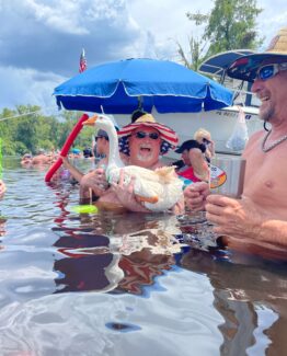 People enjoying a river party; man in a straw hat and sunglasses holds a large white swan float while others swim nearby under a blue umbrella boat backdrop on a sunny day.