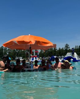 Group of people in a pool under a large orange umbrella on a sunny day photographed at a water recreation area.