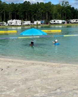Two swimmers in a lake beside a floating bright blue umbrella on a sunny day, with campers along the shore in the background.