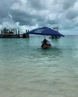 Couple embracing under a blue Tommy Bahama umbrella in shallow turquoise water near a dock.