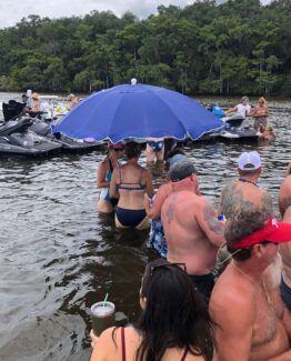 Crowded lakeside scene with swimmers in the water near parked jet skis and a large blue umbrella.