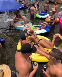 Crowded riverfront party with people in swimsuits on colorful inflatable tubes and a large blue shade umbrella nearby.