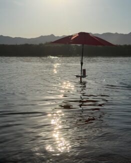 Red umbrella on a small floating platform in a sunlit lake, with sparkling water and distant hills.