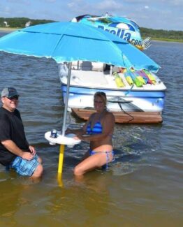 Two people stand knee-deep in shallow water beside a small anchored boat with a bright blue umbrella shade and gear on its stern.