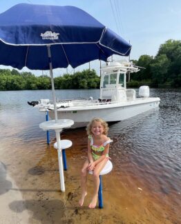 Young girl in a colorful swimsuit sits on a white stool at the water’s edge beside a white boat with a large blue canopy on a lake.