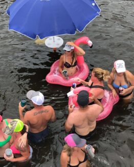 People in swimwear ride pink flamingo pool floats under a large blue umbrella, holding drinks in a crowded water party.