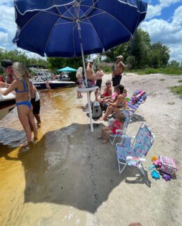 People in swimsuits enjoy a sunny lakeside beach under a large blue umbrella; boats are docked nearby and kids play in the shallow water.