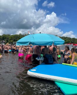 Crowded lakeshore scene with people in swimsuits standing in shallow water under a large turquoise canopy on a boat docked at the shore.