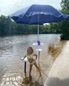 Girl in a bright swimsuit stands knee-deep in lake water under a large blue beach umbrella on a sandy lakeshore.