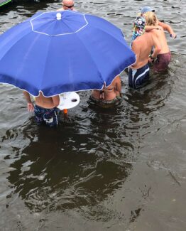 People standing in knee-deep water by a large blue beach umbrella on a dock or shoreline.