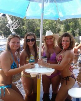 Four women in colorful bikinis posing together around a white drink table under a blue umbrella on a lake, smiling at the camera.