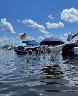 Boats docked at a sunny marina with blue umbrellas, a waving American flag, and people in the water near the vessels under a bright blue sky.