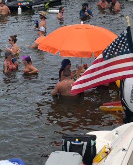 Group of people in swimwear standing and chatting in shallow water at a party, with an orange umbrella and American flag nearby.