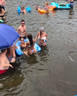 Group of people in swimwear wading in water with inflatable floats and a blue umbrella nearby at a lakeside/shore party scene.