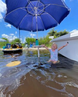 Child splashing in shallow water under a large blue patio umbrella near a white boat on a sunny day.