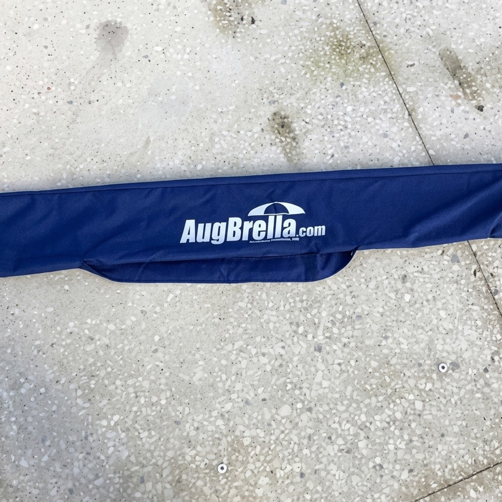 Blue fabric sleeve with the white 'AugBrella.com' logo and umbrella icon laid on a speckled concrete floor, viewed from above.