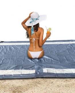 Woman in a white bikini sits on a blue beach mat on the sand, holding a citrus drink in a glass with a straw and lime slice behind her hat.