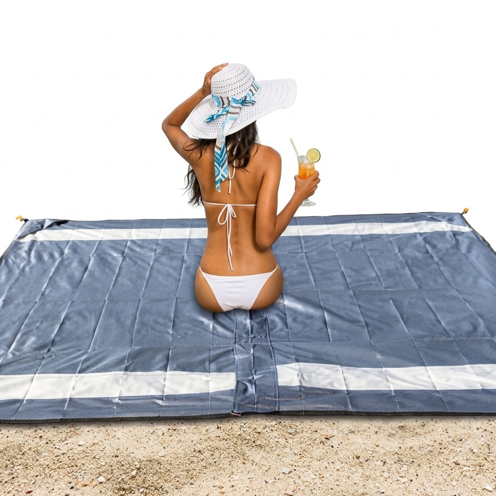 Woman in a white bikini sits on a blue beach mat on the sand, holding a citrus drink in a glass with a straw and lime slice behind her hat.