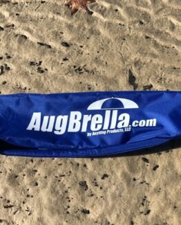 Blue fabric bag on sandy beach displaying the logo 'AugBrella.com' and 'by Anyling Products, LLC' text near the bottom edge.