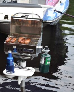 Portable stainless grill mounted on a small boat, cooking four sausages over the open lid by the water with a propane tank attached nearby