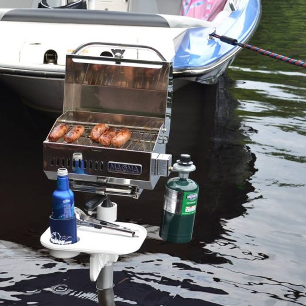 Portable stainless grill mounted on a small boat, cooking four sausages over the open lid by the water with a propane tank attached nearby
