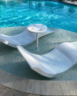 White poolside lounge chairs on a shallow pool deck with a small circular cup-holder table between them, bright blue pool water in the background.