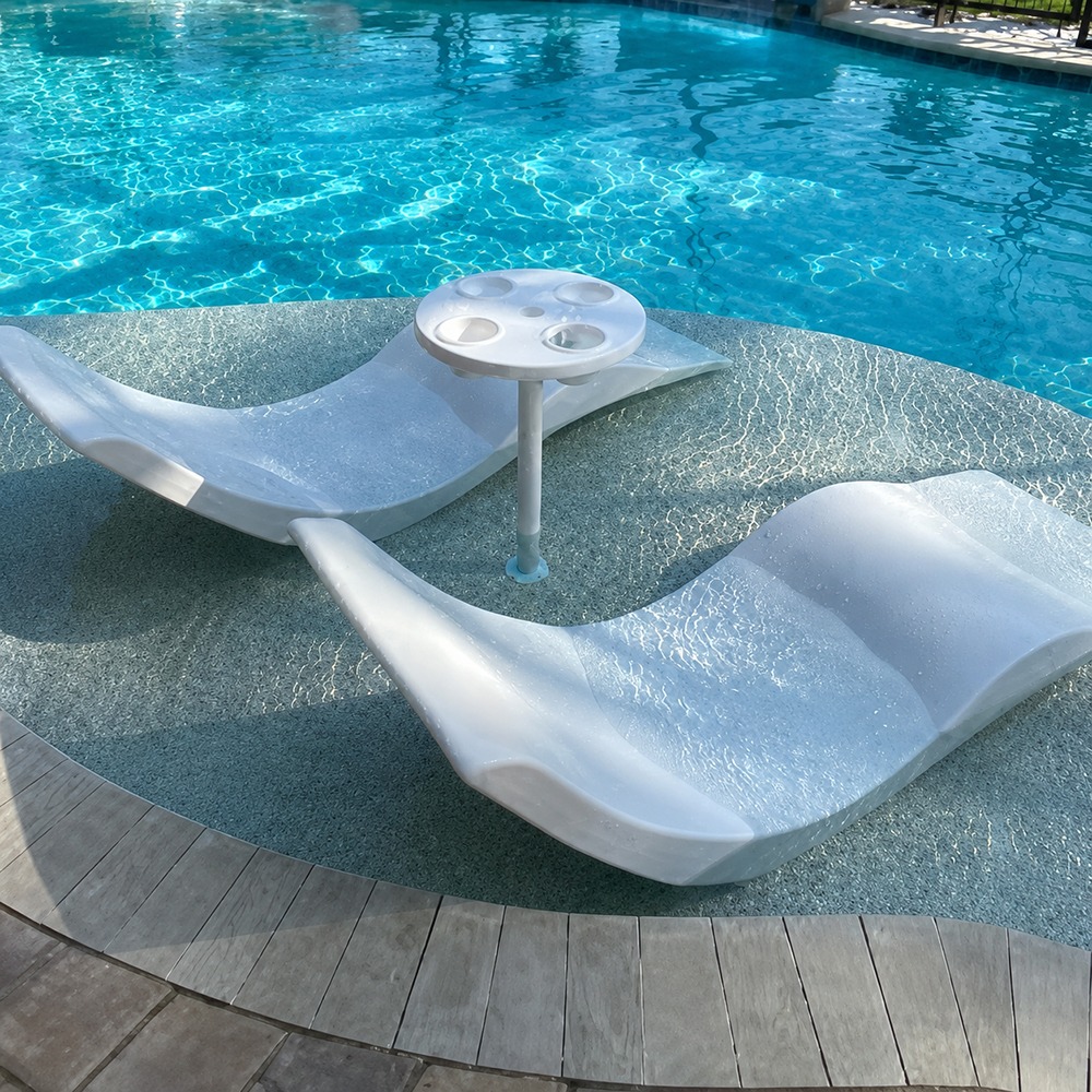 White poolside lounge chairs on a shallow pool deck with a small circular cup-holder table between them, bright blue pool water in the background.