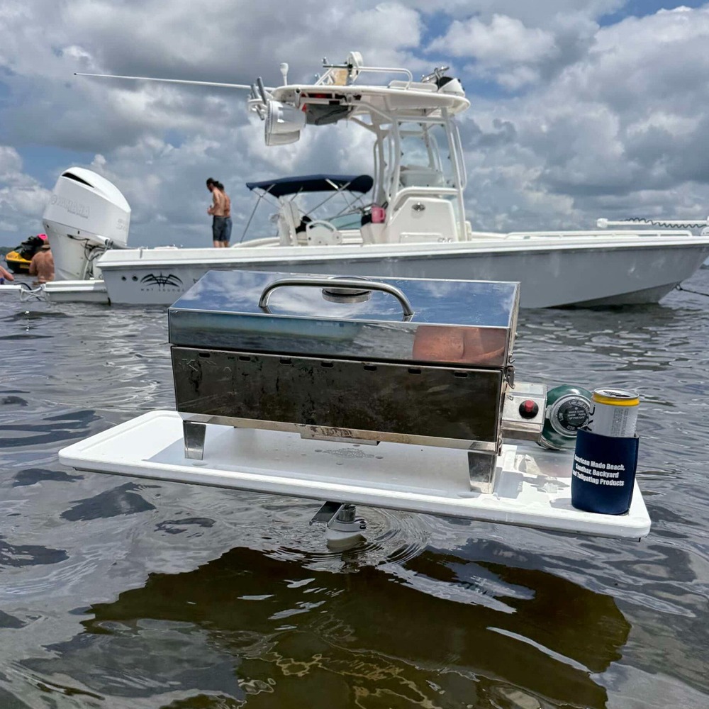 Portable stainless steel grill mounted on a white floating platform on open water, with boats and people in the background.