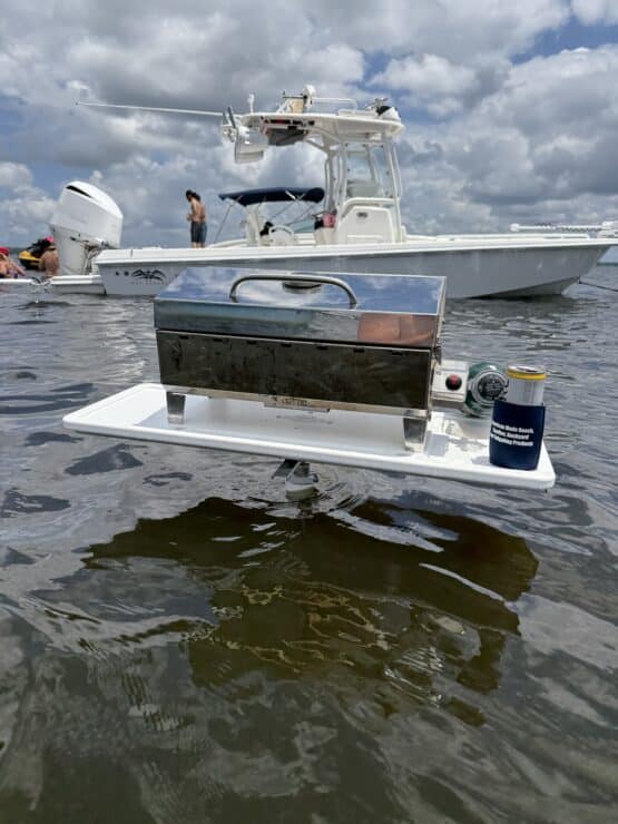 Small portable gas grill mounted on a white floating platform on the water, with a larger boat and people in the background.